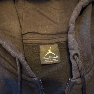 Svart hoodie från Jordan - Säljer en svart hoodie från Jordan med dragkedja och huva. Den har en vit logga på bröstet och dragsnören vid huvan. 