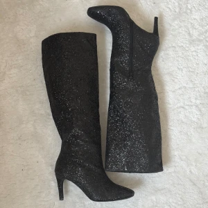 Svarta glittriga stövlar med klack - Säljer dessa ur snygga svarta glittriga klackar. Df slutar i knähöjd och perfekt för fest. Bra kvalite och inget glitter fall, har endast använt 3 gånger! De har en dragkedja på sidan för enkel på- och avtagning. Fina att ge en extra touch till din outfit. Från Nelly, men slutsålda🌷(true to size)