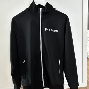 Svart track jacket från Palm Angels - Säljer en svart track jacket från Palm Angels med vita detaljer. Jackan har en dragkedja framtill och vita ränder längs ärmarna. Den är långärmad och har en hög krage. Perfekt för en sportig look. Kvitto o tags tillkommer 
