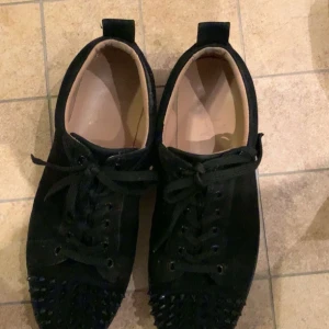 Louboutin Loubs - Snygga svarta sneakers från Louboutin med nitar på tån och klassisk röd sula. Skorna har snörning och är tillverkade i mocka för en lyxig känsla. Perfekta för att ge din stil en edgy touch.