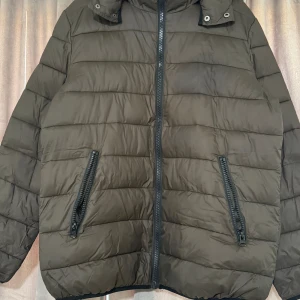 Brun quiltad jacka från Siberian - Säljer en brun quiltad jacka från Siberian med dragkedja och huva. Jackan har en pufferdesign och två praktiska fickor med dragkedjor framtill. Perfekt för kyligare dagar.