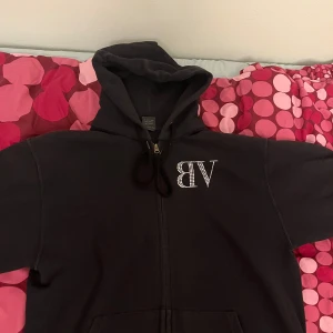 Svart hoodie med strassdetaljer från Valdi - Säljer en svart hoodie från bvaldi med strassdetaljer som bildar bokstäverna 'VB' på framsidan och 'BVALDI' på baksidan. Hoodien har dragkedja och en klassisk huva. Perfekt för en stilren och bekväm look.
