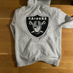 Grå Raiders hoodie från NFL - Säljer en grå hoodie med Raiders-logga från NFL. Tröjan har en stor logga på ryggen och text på framsidan. Den är tillverkad i mjukt material och har en klassisk känguruficka samt justerbar huva. Perfekt för sportfantasten!