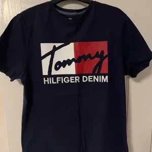Mörkblå t-shirt från Tommy Hilfiger - Snygg mörkblå t-shirt från Tommy Hilfiger med det ikoniska logotyptrycket i vitt och rött på bröstet. Perfekt för en casual look.