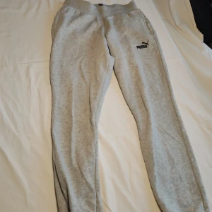 Grå mjukisbyxor från Puma - Snygga och bekväma grå mjukisbyxor från Puma. Perfekta för en avslappnad stil med elastisk midja och ribbade muddar vid bensluten. Puma-loggan är tryckt på sidan för en sportig touch.