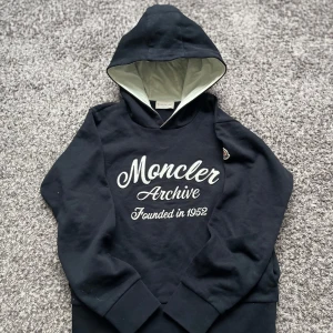 Marinblå hoodie från Moncler  - Snygg marinblå hoodie från Moncler med texten 'Moncler Archive Founded in 1952' på framsidan. Den har en huva med ljus insida och Monclers logo på armen den är Perfekt för en stilren och bekväm look.  Storlek 12 år 152