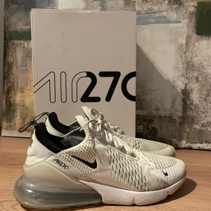 Nike Air 270 i färgen vit  - Snygga Nike Air 270 sneakers i vitt med svarta detaljer. Skorna har en stor synlig luftkudde i hälen för extra komfort och stil. Ovandelen är i ventilerande meshmaterial med snörning för bra passform. Perfekta för en sportig look. Nike märket där fram på höger skon är lite av men går att limma fast igen⭐️
