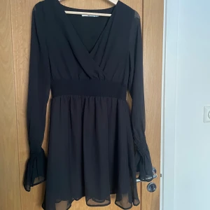 Svart klänning från NA-KD - Elegant svart klänning från NA-KD med omlott-design och långa ärmar. Klänningen har en smickrande v-ringning och elastisk midja för en bekväm passform. Perfekt för en stilren look.