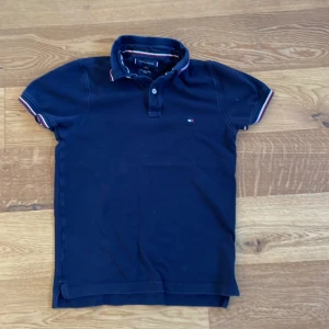 Mörkblå pikétröja från Tommy Hilfiger - Snygg mörkblå pikétröja från Tommy Hilfiger i ekologisk bomull. Tröjan har en slim fit passform och korta ärmar med röd och vit randdetalj vid ärmslut och krage. Perfekt för en stilren look!