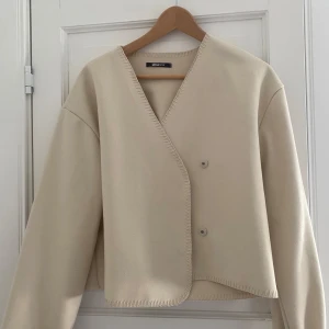 Beige omlottjacka från Gina Tricot - Snygg beige omlottkappa från Gina Tricot med v-ringad design och knappar framtill. Jackan har långa ärmar och är lite tunnare som passar perfekt för både höst och vår. Använd fåtal gånger och finns inga defekter, skicket är som ny. 