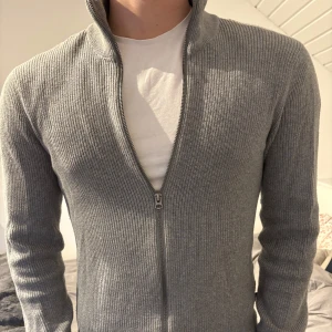 Grå ribbad tröja med dragkedja från Dobber - Säljer en stilren grå ribbad tröja från Dobber. Tröjan har en dragkedja framtill och långa ärmar, perfekt för en avslappnad look. Den är i storlek small och passar bra till både vardag och mer uppklädda tillfällen.