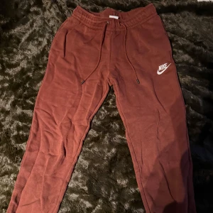Bruna mjukisbyxor från Nike - Säljer ett par bruna mjukisbyxor från Nike med dragsko i midjan och en vit logga på benet. Perfekta för en avslappnad stil.