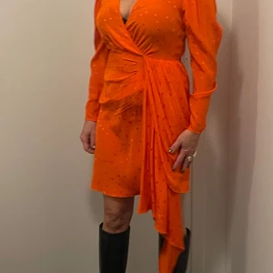 Orangeröd omlottklänning från Essentiel Antwerp. - Snygg orangeröd omlottklänning från Essentiel Antwerp med prickigt mönster. Klänningen har långa ärmar och en v-ringad design. Perfekt för att sticka ut med sin livfulla färg och stiliga detaljer.