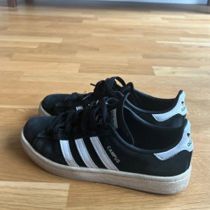 Svarta Adidas Campus sneakers - Säljer ett par svarta Adidas Campus sneakers med vita detaljer och klassiska tre ränder på sidan. Skorna har en vit sula och snörning framtill. Perfekta för en avslappnad stil.