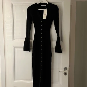 Svart långklänning från Adoore - Elegant svart långklänning från Adoore med ribbad struktur och knappar längs hela framsidan. Klänningen har en v-ringad design och långa ärmar, perfekt för en stilren look.