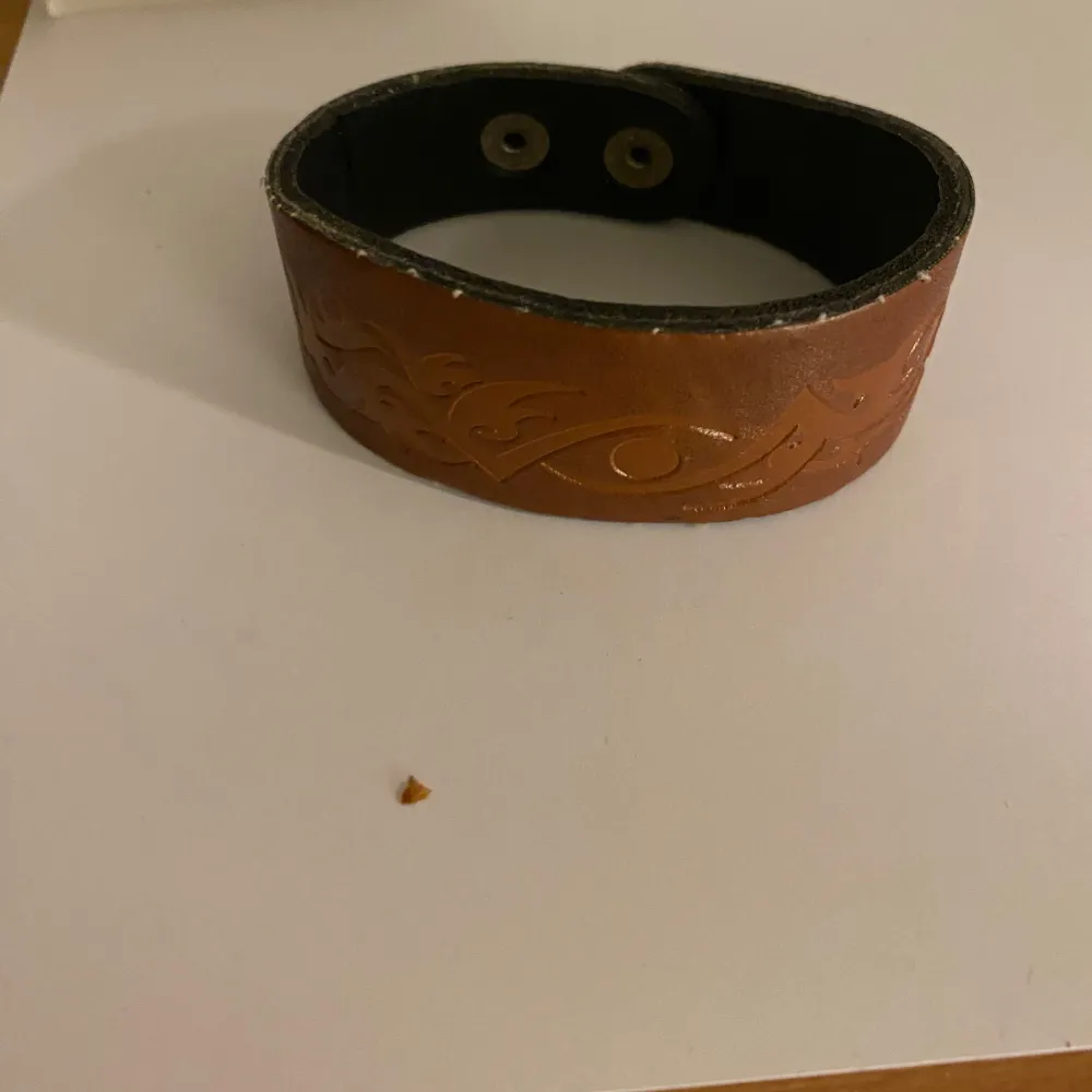Säljer ett snyggt brunt läderarmband med ingraverat mönster. Armbandet har en bredd på cirka 5 cm och är perfekt för att ge en stilren touch till din outfit. Det har en enkel knäppning med tryckknappar.. Asusteet.