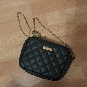 Svart quiltad axelväska med kedja - Snygg svart axelväska i quiltat mönster med guldfärgad kedja som axelrem. Perfekt för att ge en elegant touch till din outfit. Väskan har en dragkedja för säker förvaring.