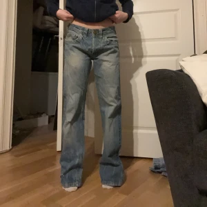 Blå jeansbyxor från rodi denims - Säljer ett par blå jeansbyxor från rodi denims med en straight passform. Byxorna har en knappgylf och är dekorerade med slitningar för en cool look. Perfekta för en avslappnad stil.