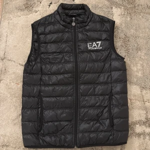 Svart dunväst från Emporio Armani - Snygg svart dunväst från Emporio Armani med EA7-logga på bröstet. Västen har en dragkedja framtill och är quiltad för extra stil och värme. Perfekt för kyligare dagar när du vill hålla stilen.