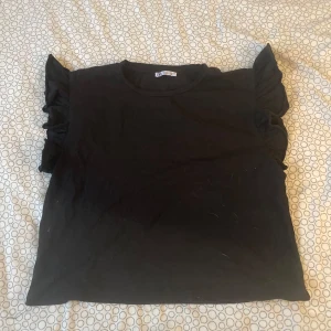 Svart topp med volangärmar från Zara - Snygg svart topp från Zara med volangdetaljer på ärmarna. Perfekt för att ge en extra touch till din outfit. Tillverkad i ett mjukt och bekvämt material. Köpte för några år sedan. Använd ett fåtal gånger.