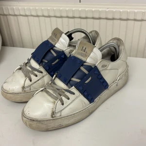 Velentino skor - Snygga vita sneakers från Valentino med blå detaljer och nitar. Skorna har en unik design med snörning och en robust sula. Perfekta för att ge en cool touch till din outfit. Skick 7/10 saknar 3 nitar