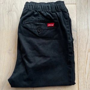 Svarta chinos från Levi's - Snygga svarta chinos från Levi's med elastisk midja och snörning. Byxorna har en tapered passform och är perfekta för en stilren look. De har både fram- och bakfickor med knappdetalj på baksidan. Storlek 158-164. Fläckar syns på bild 3.