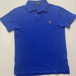 Blå pikétröja från Ralph lauren.  - Säljer en snygg blå pikétröja från Ralph Lauren i slim fit. Tröjan har korta ärmar och en klassisk krage med knappar. Den ikoniska loggan är broderad i orange på bröstet. Perfekt för en stilren look! Storlek: M (S i verkligheten) Den är knappt använd. Skick :10/10
