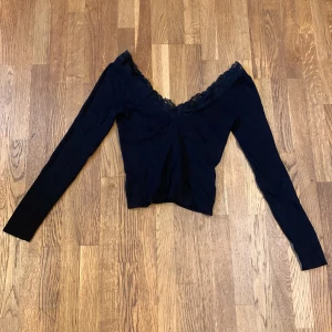 Svart ribbad topp från H&M Divided - Säljer en svart ribbad topp från H&M Divided med spetsdetaljer vid den v-ringade halsen. Toppen har långa ärmar och en croppad passform.