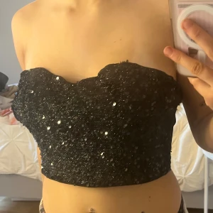 Svart glittrig tubtopp - Säljer en snygg svart tubtopp med glittrande paljetter. Perfekt för en kväll ute eller fest. Toppen är axelbandslös och har en tight passform som framhäver figuren.