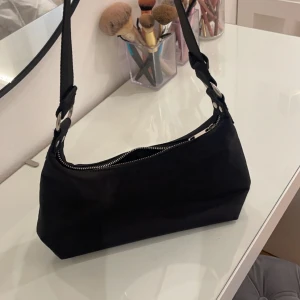 Svart axelväska med dragkedja - Snygg svart axelväska i syntetmaterial med en praktisk dragkedja upptill. Perfekt för att bära dina nödvändigheter med stil. Enkel och elegant design som passar till många olika outfits.