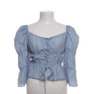 Blå blus från Stradivarius - Säljer en söt blå blus från Stradivarius med långa ärmar och knytning framtill. Blusen har en lätt rynkad design och volangdetaljer vid nederkanten. Bra skick förutom att den kanske ser lite skrynklig ut!