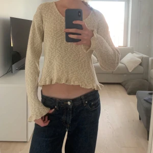 Beige ribbad tröja - Säljer en beige ribbad tröja med v-ringning och långa ärmar. Tröjan är croppad med volangdetaljer vid nederkanten och ärmsluten. Perfekt för en avslappnad stil.