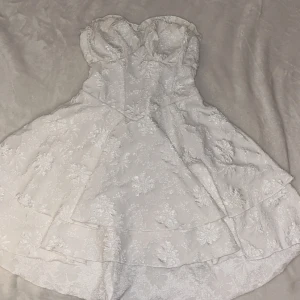 Vit axelbandslös klänning med blommigt mönster - Charmig vit axelbandslös klänning som formar midjan med ett vackert blommigt mönster. Klänningen har en volangdetalj Den har en kort längd och en elegant passform som framhäver figuren. Bust:74-100 cm, Waist Size:67-96 cm, Length:64 cm