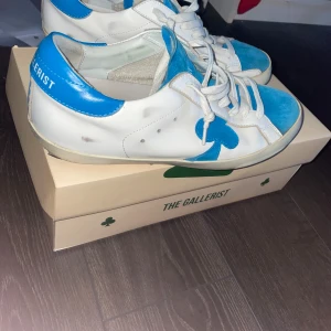 Vita och blå sneakers från The Gallerist - Snygga vita sneakers med blå detaljer från The Gallerist. Skorna har en unik blå klöverdesign på sidan och matchande blå häl och tåparti. De är utrustade med vita snören och har en klassisk sneakerstil. Finns två hål i sulan som ni ser på bilden