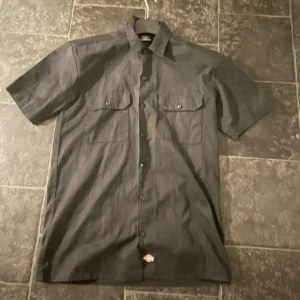 Svart kortärmad skjorta från Dickies - Snygg svart kortärmad skjorta från Dickies med klassisk krage och knappar framtill. Skjortan har två bröstfickor med lock och knappstängning. Känns mer som en Medium
