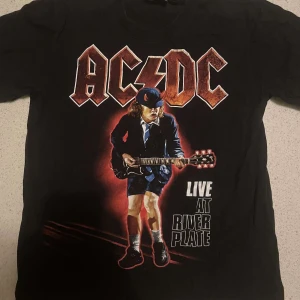 Svart AC/DC t-shirt - Cool svart t-shirt med AC/DC-tryck fram och bak. Framsidan har ett stort tryck av en gitarrist och texten 'Live at River Plate'. Perfekt för rockälskare! 
