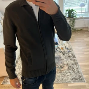 Dressman cardigan  - Säljer en stilren mörk grön cardigan med dragkedja. Cardiganen har långa ärmar och ribbade detaljer vid ärmslut och nederkant. Perfekt för en casual look.