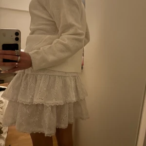 Vit volangkjol - Charmig vit kortkjol med volanger och broderade detaljer. Perfekt för en söt och romantisk stil. Kjolen har ett lager av spets som ger en extra touch av elegans.