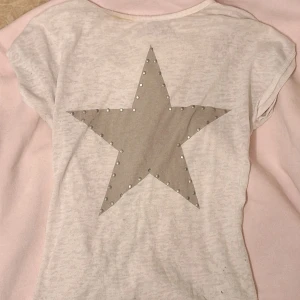 Vit topp med stjärna - Superfin kortärmad topp med en grå stjärna på framsidan, dekorerad med små nitar. Toppen är gjord i ett mjukt bomullsmaterial & endast lite genomskinlig (så det blir som ett slags snyggt mönster) 🫶🏼 endast en defekt på kragen men ska gå bort I tvätten! använd sparsamt så annars i bra skick och färgen är ljusare i verkligheten.💓