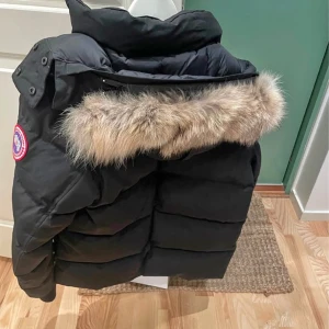 Canada goose  - Säljer en svart dunjacka från Canada Goose med avtagbar pälsdetalj på huvan. Jackan har en klassisk design med dragkedja och knappar framtill samt långa ärmar. Perfekt för kalla vinterdagar.