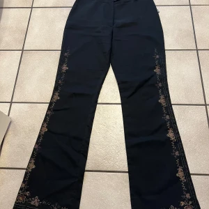 Svarta byxor med broderade detaljer - Eleganta svarta byxor med vackra broderade blommor längs sidorna. Byxorna har en rak passform och hög midja, vilket ger en stilren look. Perfekta för att lägga till en touch av elegans till din outfit. Står storlek L, men tycker att den är S/M, midja 68 cm. Använda en gång.