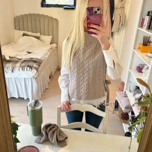 Beige stickad väst med knytband - Säljer en snygg beige stickad väst med flätmönster och knytband i sidorna. Västen har en hög krage och är perfekt för lager-på-lager-stil. Passar bra till både skjorta och t-shirt. Har matchande torghandskar,(4 och 5 bilden).