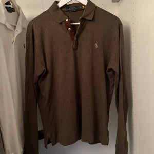 Brun långärmad pikétröja från Ralph Lauren - Säljer en brun långärmad pikétröja från Ralph Lauren i custom slim fit. Tröjan har en klassisk krage och knappar framtill. Den är dekorerad med det ikoniska broderade polospelar-emblemet på bröstet. Perfekt för en stilren look.