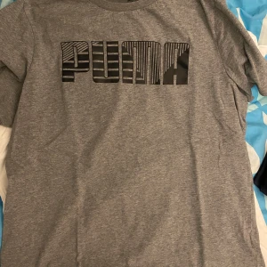 Grå t-shirt från Puma - Säljer en grå t-shirt från Puma med ett coolt mönstrat tryck på bröstet. T-shirten är kortärmad och har en klassisk rund halsringning. Perfekt för en avslappnad stil. Inga difekter och fläckar!