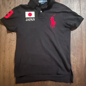 Svart pikétröja från Ralph Lauren - Snygg svart pikétröja från Ralph Lauren med röd logga och nummerdetalj på ärmen. Tröjan har en japansk flagga och texten 'Japan' på bröstet. Perfekt för en stilren look.