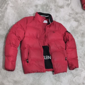 Röd pufferjacka från Calvin Klein - Säljer en snygg röd pufferjacka från Calvin Klein i storlek S, för super bra pris då jag använder en annan jacka. I ok skick. Värd över 2000 kr i butik. Perfekt för kyligare dagar eller regn. Små defekter i form av lätt slitage på framsidan och på ärmen på baksidan. Se bild.