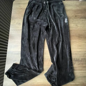 Svarta mjukisbyxor från Juicy Couture - Snygga svarta mjukisbyxor från Juicy Couture i mjukt sammetstyg. Byxorna har en elastisk midja med snörning och dragkedjefickor. Dekorerade med glittrande stenar som formar ordet 'Juicy' på baksidan och en logotyp på framsidan. Många stenar har trillat av från trycket på baksidan