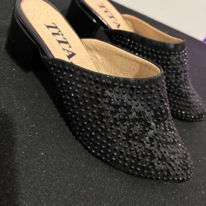 Svarta slip-in skor med pärlor - Eleganta svarta slip-in skor från Tita med glittrande pärlor och blommönster. Skorna har en låg klack och en spetsig tå, vilket ger en stilren look. 