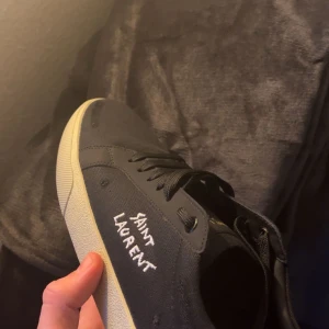 Svarta sneakers från Saint Laurent - Snygga svarta sneakers från Saint Laurent med vit sula och handskriven logga på sidan. Skorna har snörning och är tillverkade i ett slitstarkt material. Perfekta för en stilren look. Priset kan diskuteras!!