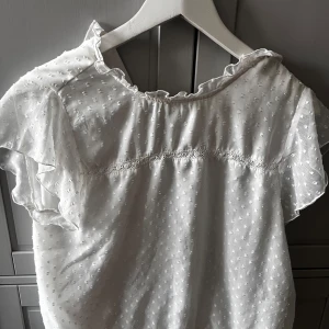 Vit blus med volangdetaljer -  vit blus med volangdetaljer och ett diskret prickigt mönster. Blusen har korta ärmar och en rund halsringning med små volanger.  Sitter som M/S